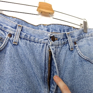Op de afbeelding: Lichtblauwe spijkerbroek met een klassiek ontwerp met vijf zakken. De jeans heeft een knoop- en ritssluiting, riemlussen en een kleine bruine leren patch. De jeans hangt aan een metalen en houten hanger.