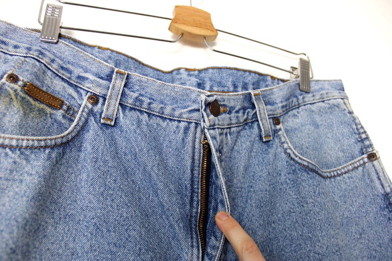 Op de afbeelding: Lichtblauwe spijkerbroek met een klassiek ontwerp met vijf zakken. De jeans heeft een knoop- en ritssluiting, riemlussen en een kleine bruine leren patch. De jeans hangt aan een metalen en houten hanger.
