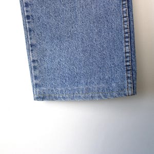 Op de afbeelding: Detail van de zoom en de zijnaad van een lichtblauwe denim jeans. De jeans heeft een klassiek vijf-zakken ontwerp en is gemaakt van een duurzame denim stof. De stiksels zijn zichtbaar.