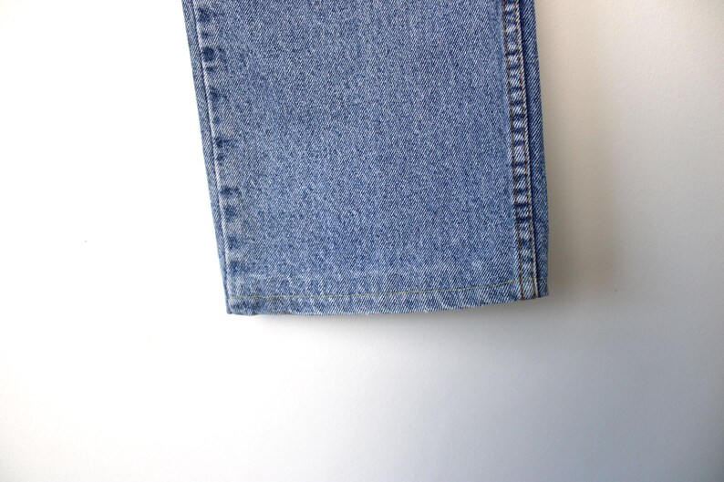 Op de afbeelding: Detail van de zoom en de zijnaad van een lichtblauwe denim jeans. De jeans heeft een klassiek vijf-zakken ontwerp en is gemaakt van een duurzame denim stof. De stiksels zijn zichtbaar.