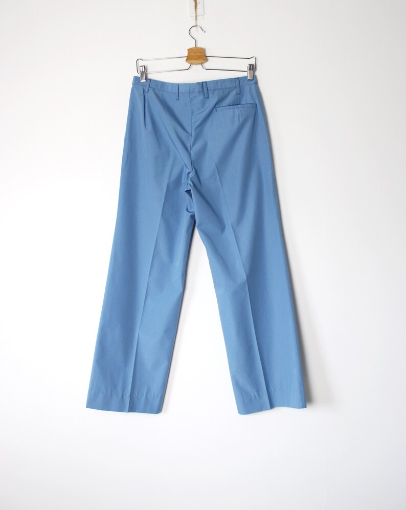 K&ouml;nnte beinhalten: Eine hellblaue, weite Hose, die an einem Holzb&uuml;gel an einer wei&szlig;en Wand h&auml;ngt.
