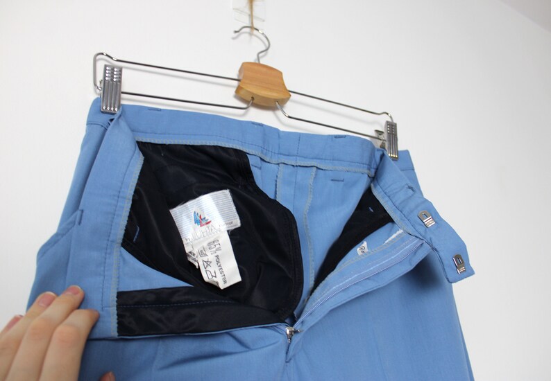K&ouml;nnte beinhalten: Eine hellblaue Hose mit schwarzem Bund und Rei&szlig;verschluss. Die Hose hat ein Etikett mit der Aufschrift "65% Polyester & 35% Baumwolle" und ein kleines Logo mit einem Vogel.