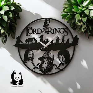 May include: Black metal wall clock with a circular design featuring the text "The Lord of the Rings" and silhouettes of characters from the movie. The clock has a white clock face with black hands.