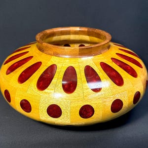 Puede incluir: Un cuenco de madera hecho a mano con un diseño amarillo y rojo. El cuenco presenta una base de madera de color claro con una serie de incrustaciones rojas ovaladas y circulares. El borde es de madera más oscura, creando un contraste. Este cuenco decorativo es una obra de arte única.