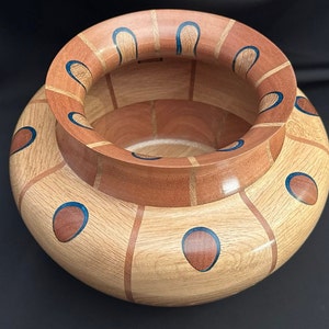 Peut inclure: Un bol en bois fait à la main avec une base large et arrondie et un bord évasé. Le bol présente un bois de couleur claire avec des accents de bois plus foncés et des détails bleus incrustés. Le design est géométrique et décoratif.