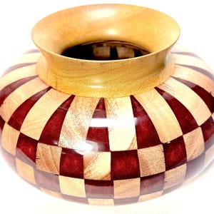 Puede incluir: Un jarrón de madera hecho a mano con un cuerpo ancho y redondeado y un borde acampanado. El jarrón presenta un patrón de tablero de ajedrez de madera de color marrón rojizo y claro, creando un contraste visual llamativo. El borde es de un tono más claro.
