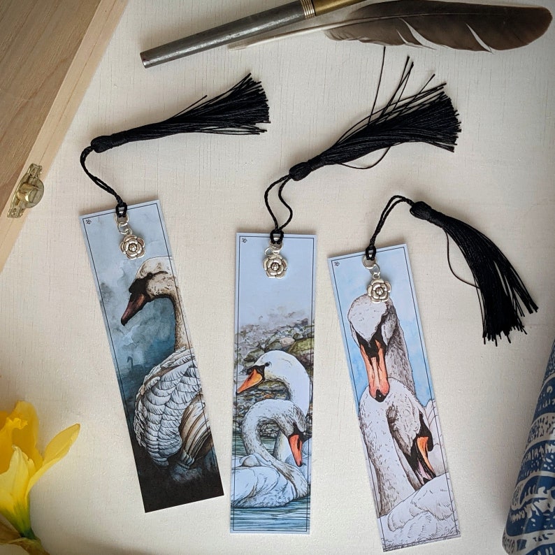 May include: Three bookmarks with black tassels and silver charms featuring a floral design. Each bookmark features a different illustration of swans in a watercolor style.