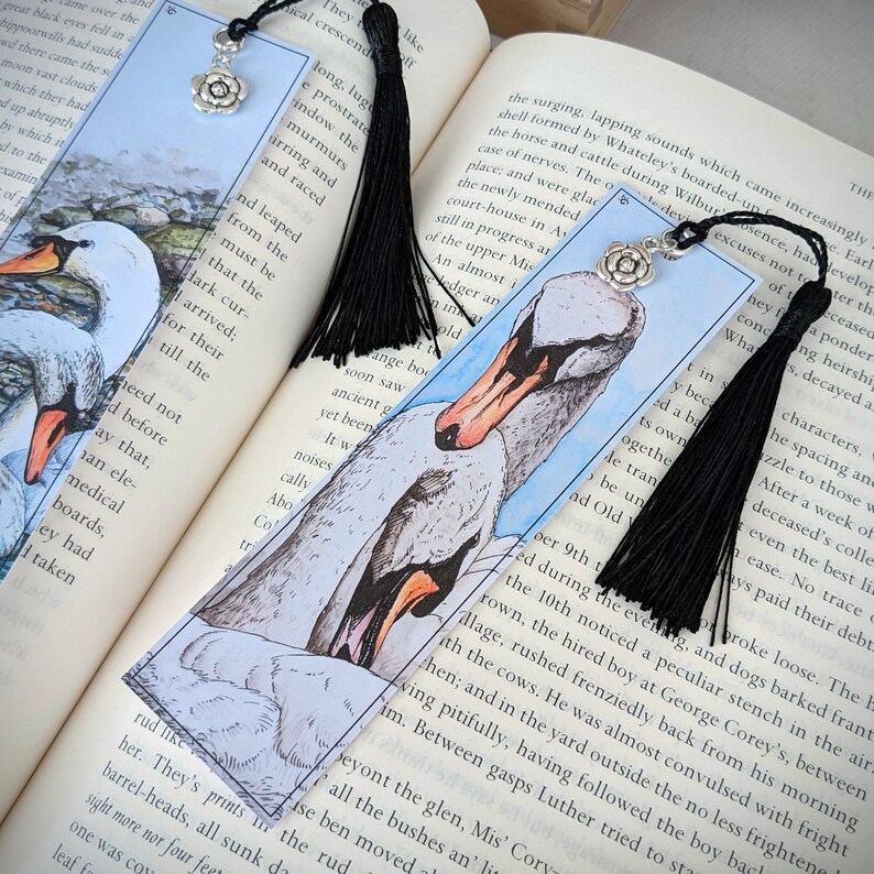 May include: Two black tassel bookmarks with silver flower charms. One bookmark features a watercolor illustration of two swans on a blue background. The other bookmark features a watercolor illustration of a swan on a blue background. The text "What they need not be told before that, any ele- medical boards, they had taken"