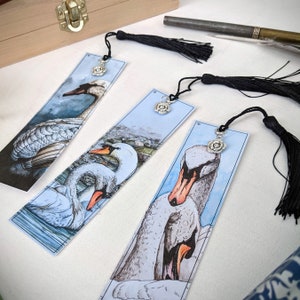 May include: Three bookmarks featuring watercolor illustrations of swans on a blue background. Each bookmark has a silver charm in the shape of a rose.