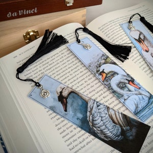 May include: Two white swans with black eyes and beaks are depicted on a blue and white watercolor bookmark. The bookmark has a black tassel and a silver charm in the shape of a flower.