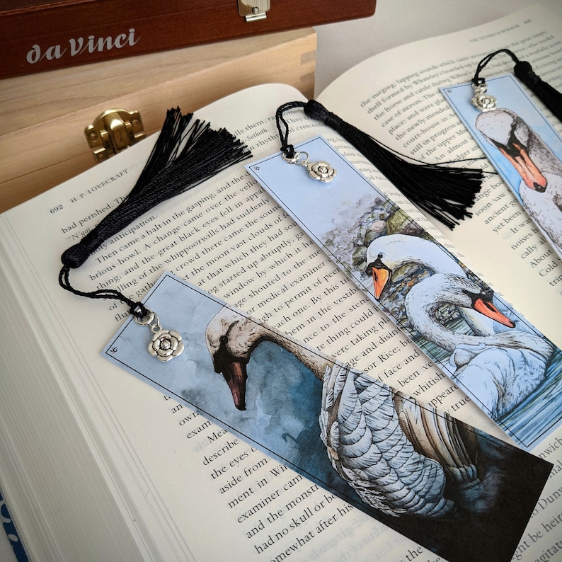 May include: Two white swans with black eyes and beaks are depicted on a blue and white watercolor bookmark. The bookmark has a black tassel and a silver charm in the shape of a flower.