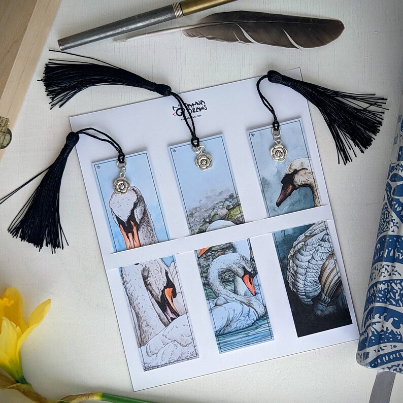 May include: Six white swan bookmark designs with silver charms. Each bookmark features a different swan design, including a swan with a flower charm, a swan with a crown charm, and a swan with a heart charm. The bookmarks are printed on white cardstock and have black ribbon ties.