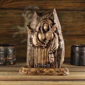May include: A carved wooden statue of a robed figure holding a feather and a mask. The statue is in shades of brown and beige, with the inscription "N<mxln" at the base. Two small, dark containers flank the statue.