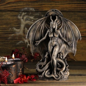 May include: A dark gray, winged, female figurine with horns and a snake at her feet. The figurine is standing on a wooden surface with a candle in the foreground.