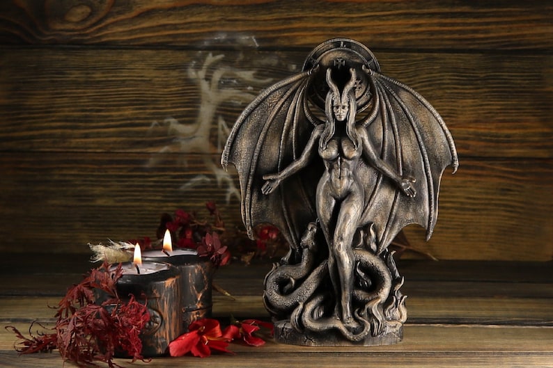 May include: A dark gray, winged, female figurine with horns and a snake at her feet. The figurine is standing on a wooden surface with a candle in the foreground.