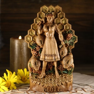 May include: A wooden figurine of a woman standing in front of a honeycomb. She is holding a honeycomb in her right hand and a flower in her left hand. The figurine is painted in shades of brown and gold. The base of the figurine is inscribed with the word "BEOLIE".