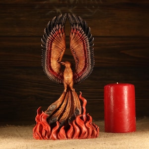 Op de afbeelding: Een gedetailleerde sculptuur van een feniks met uitgespreide vleugels, in tinten bruin, rood en zwart, oprijzend uit gestileerde vlammen. Een rode pilaar kaars staat naast de sculptuur. De achtergrond is donker hout.