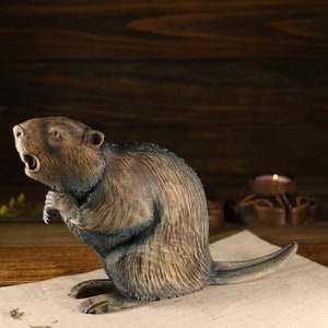 May include: A wooden figurine of a beaver with a distressed brown and blue paint finish. The beaver is standing on its hind legs with its front paws raised. The figurine is on a light brown surface.