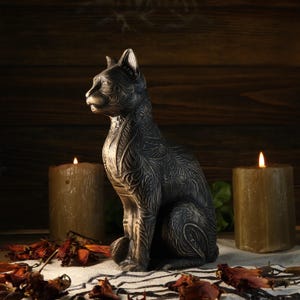 Può includere: Una statua di gatto grigio scuro con intricati motivi floreali è posta su un tessuto a righe bianche e nere. Due candele accese affiancano la statua e foglie secche sono sparse, creando un'atmosfera mistica.