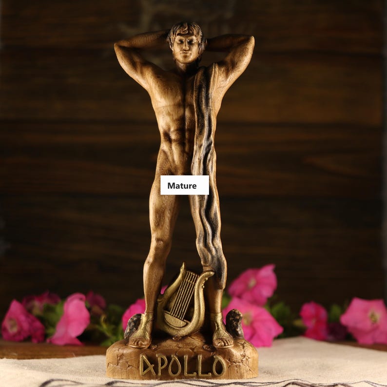 Hand-carved wooden statue of the Greek god Apollo standing in a classic athletic pose with hands behind his head. A dark draped fabric hangs over his shoulder. A golden lyre rests between his feet on a textured base with the name APOLLO. Dark rustic wooden background with pink flowers.