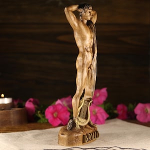 Profile view of a hand-carved wooden statue of the Greek god Apollo, captured from a side angle. The figure stands in an athletic posture with hands placed behind his head. The detailed carving of the vertical draped fabric is visible along his body. A golden lyre rests at the bottom on a rocky base. Dark wooden backdrop with pink flowers.