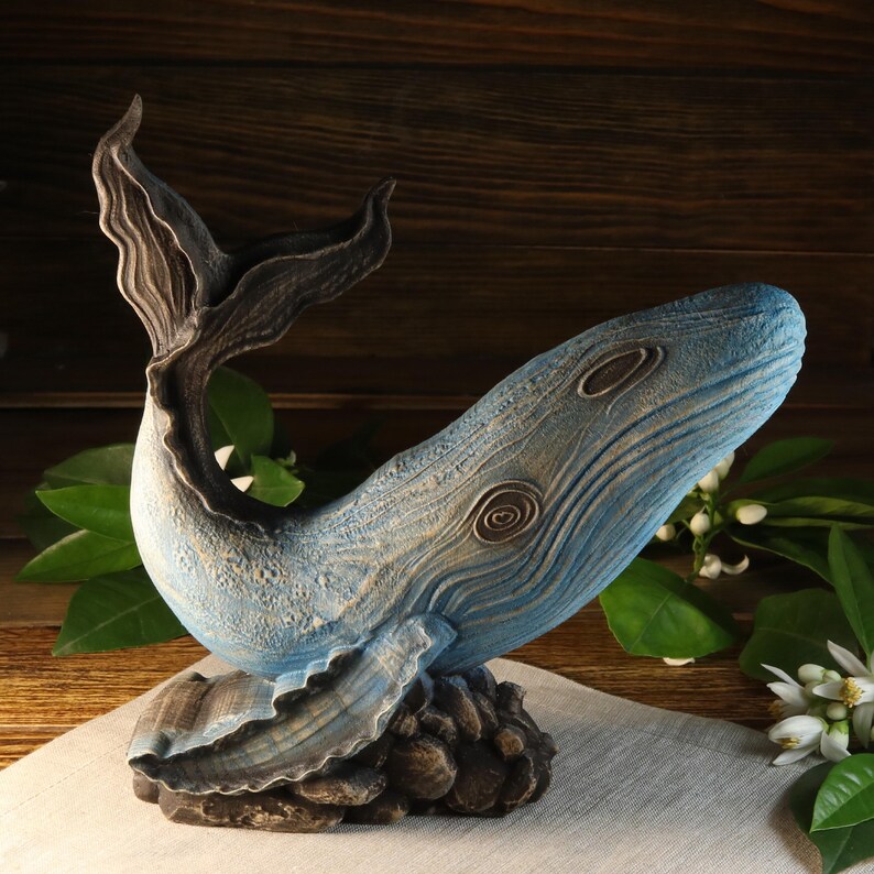 A blue and gray whale sculpture with a textured finish. The whale is resting on a rocky base. The tail is raised, and the body has detailed carvings. The sculpture is a decorative item.