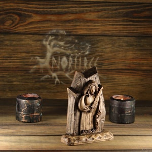 May include: A wooden sculpture of a robed figure holding a staff, with the inscription "Nsmxln" at the base. Two small, dark candle holders flank the sculpture. The background is a wooden wall with the word "NORHICE" visible.