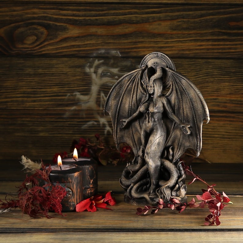 May include: A dark brown, winged demon statue with a female figure standing on a snake. The statue is on a wooden surface with two black candles.