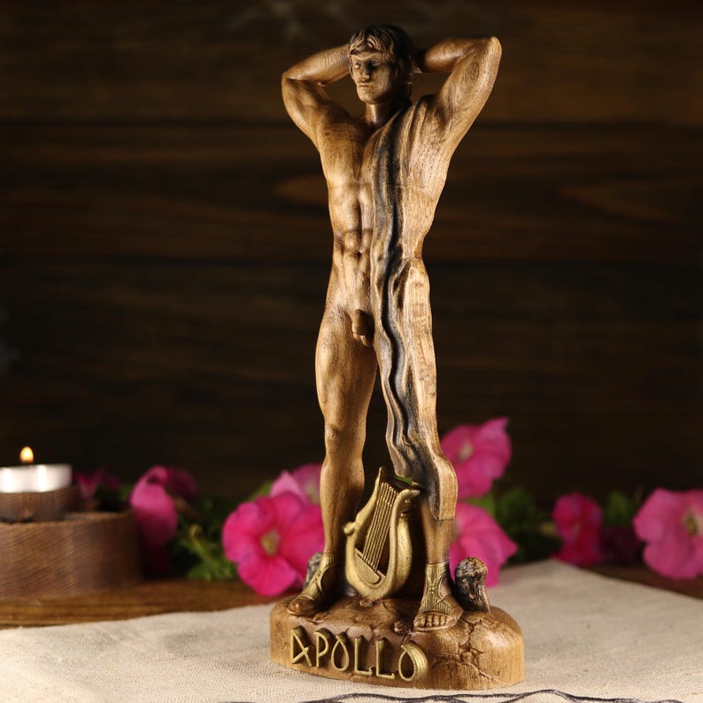 Side angle view of a hand-carved wooden statue of the Greek god Apollo standing in a classical pose with hands behind his head. The perspective showcases the texture of the carved timber and the drape of the fabric over his shoulder. A golden lyre rests near his feet on a rocky base with the name APOLLO visible. Dark rustic wood background with blurry pink flowers.