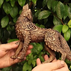 Könnte beinhalten: Eine bronzefarbene Wolfsfigur in heulender Pose, verziert mit keltischen Knotenmustern. Die Skulptur wird in Händen vor einem Hintergrund aus grünem Laub gehalten und zeigt aufwendige Details und eine verwitterte Oberfläche.