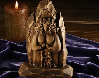 Puede incluir: Una estatua de madera tallada de una mujer con las manos juntas en oración, sobre un fondo de una estructura montañosa. La estatua es marrón y se asienta sobre una base geométrica. Una vela marrón encendida está en el fondo.