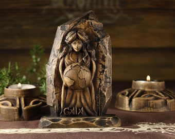 Puede incluir: Una escultura de madera de una mujer sosteniendo un globo terráqueo, con la palabra "GAIA" tallada debajo. La escultura está sobre una base de madera y está rodeada por dos candelabros.
