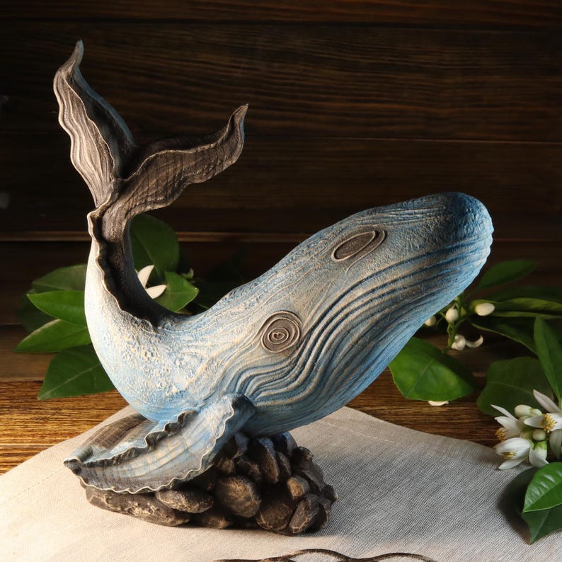 A side view of a hand-carved wood blue whale figurine, showcasing detailed ocean decor sculpture with visible wood carving textures. The sea animal sculpture is available with custom engraving options for personalization, set against a rustic background with green leaves.