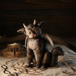 Puede incluir: Una figura de gato de fantasía marrón y beige con ojos amarillos, cuernos pequeños y alas. El gato tiene una cola larga y está sentado sobre una superficie estampada. Un pequeño candelabro está en el fondo.