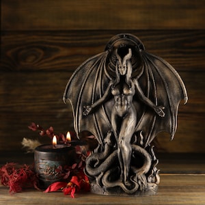 May include: A dark bronze statue of a female figure with wings and horns, standing on a snake. The statue is on a wooden surface with red flowers.