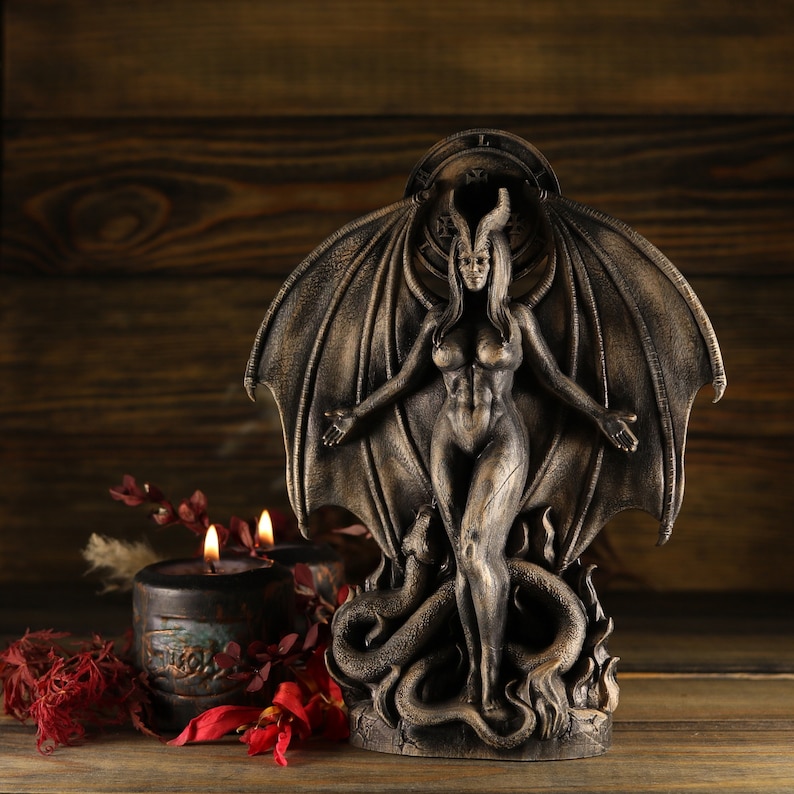 May include: A dark bronze statue of a female figure with wings and horns, standing on a snake. The statue is on a wooden surface with red flowers.