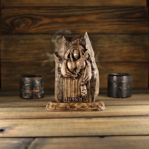 May include: A carved wooden statue of a figure holding a feather and a mask, with the word "NEMXIN" at the base. The statue is brown and stands on a wooden surface. Two small, dark brown cylindrical objects are on either side of the statue.
