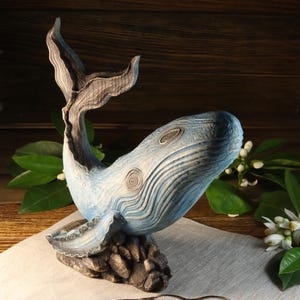 A side view of the hand-carved wood blue whale figurine, featuring a textured light blue body with dark blue accents, detailed fins, and tail. The ocean decor sculpture rests on a dark textured base and is available with a personalized engraving option, set against a wooden background.