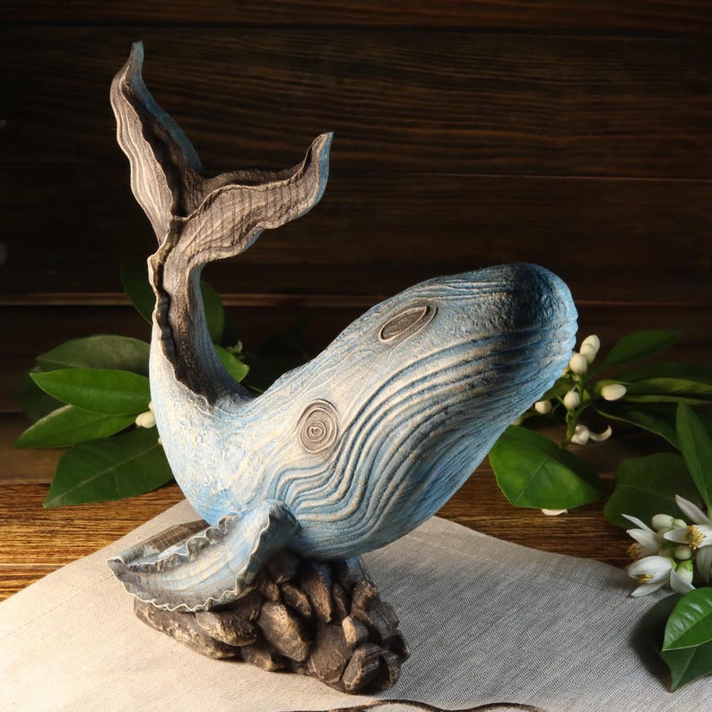A side view of the hand-carved wood blue whale figurine, featuring a textured light blue body with dark blue accents, detailed fins, and tail. The ocean decor sculpture rests on a dark textured base and is available with a personalized engraving option, set against a wooden background.