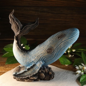 A side view of the hand-carved wood blue whale figurine from the listing, showcasing detailed ocean decor sculpture with a textured surface, blue accents, and a dark base. This sea animal sculpture is available with custom engraving options for personalization, set against a wooden background.