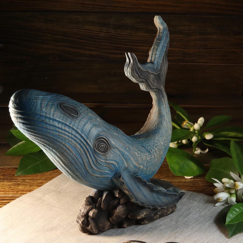 A left-side, slightly angled view of the hand-carved wood blue whale figurine. The ocean decor sculpture features intricate blue textures, detailed fins, and a high arched tail resting on a dark base. This sea animal figurine can be customized with personalized engraving.