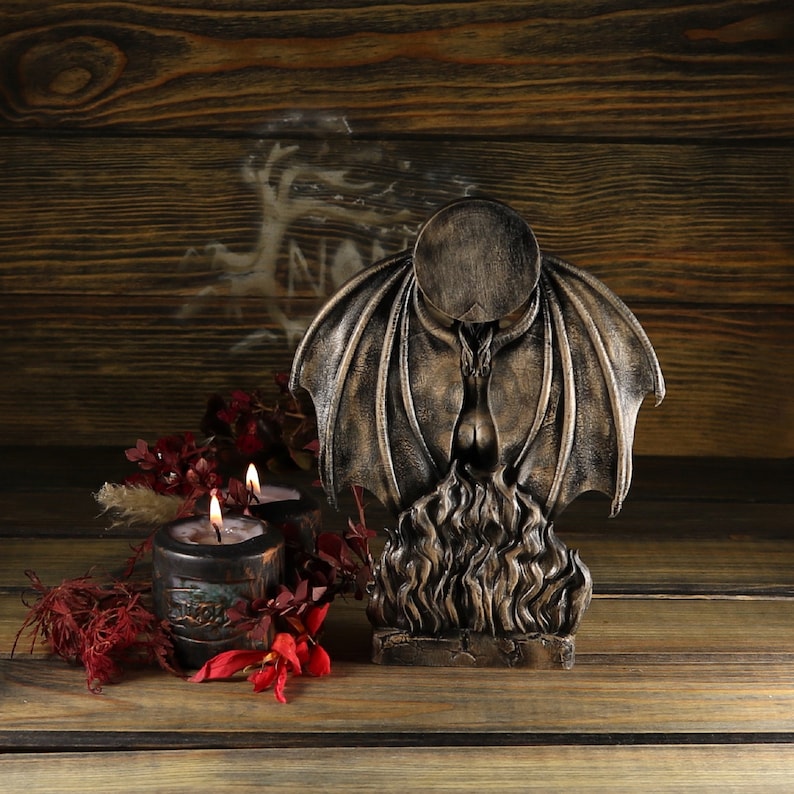 May include: A dark, gothic-style figurine of a winged creature with flames at its base. The figurine is made of a dark, textured material and has a distressed finish. Two lit candles are in front of the figurine.