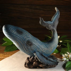 A left-side view of the hand-carved wood blue whale figurine, showing the detailed ocean decor sculpture facing left with its tail raised high. The textured sea animal sculpture rests on a dark rock-like base and is available with a personalized engraving option, set against a rustic background.