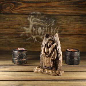 May include: A wooden carved statue of a robed figure holding a feather and a mask, with the text "Nornir" at the base. Two small candle holders with lit candles flank the statue. The background is a wooden surface.