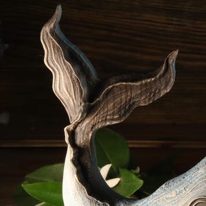 A detailed sculpture of a whale's tail, featuring a textured, dark gray finish with lighter gray highlights. The tail curves gracefully, with visible carved details. The sculpture is set against a dark wooden background, with green leaves visible.