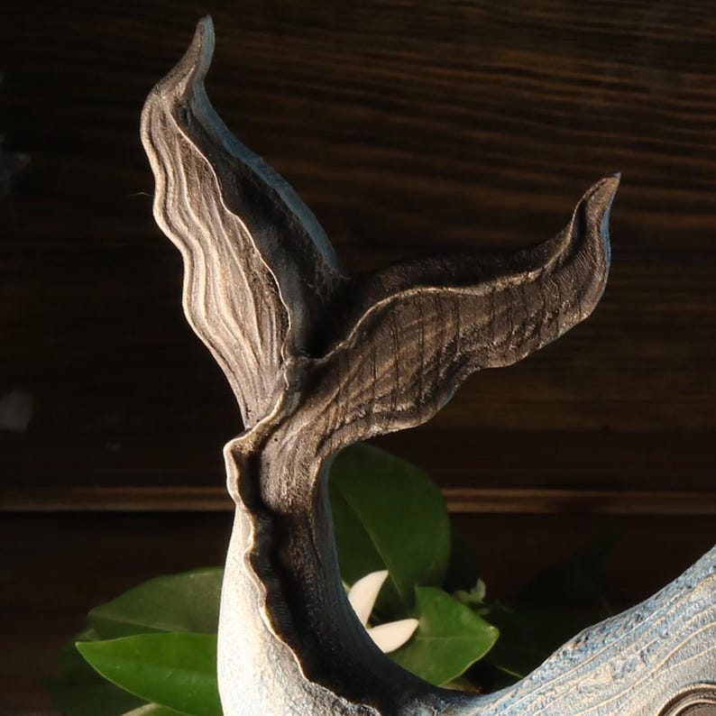 A detailed sculpture of a whale's tail, featuring a textured, dark gray finish with lighter gray highlights. The tail curves gracefully, with visible carved details. The sculpture is set against a dark wooden background, with green leaves visible.