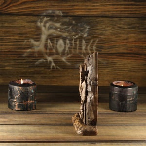 May include: A carved wooden sculpture of a figure stands on a wooden surface, flanked by two lit candles in dark ceramic holders. The background features a wooden wall with a tree and the word "NORRICE" faintly visible.