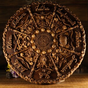Peut inclure: Une roue en bois avec des sculptures repr&eacute;sentant les huit sabbats de la Roue de l'Ann&eacute;e. La roue est brune et a une bordure brun fonc&eacute;. Les sabbats sont &eacute;tiquet&eacute;s dans le sens des aiguilles d'une montre&nbsp;: Beltane, Litha, Lughnasadh, Mabon, Samhain, Yule, Imbolc et Ostara.