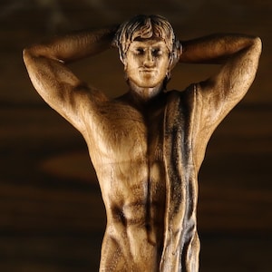 Close-up of a hand-carved wooden statue of the Greek god Apollo. The shot focuses on the detailed facial features, athletic torso, and natural wood grain of the timber. The figure has both hands behind his head with a dark draped fabric over his shoulder. Dark rustic wooden background.