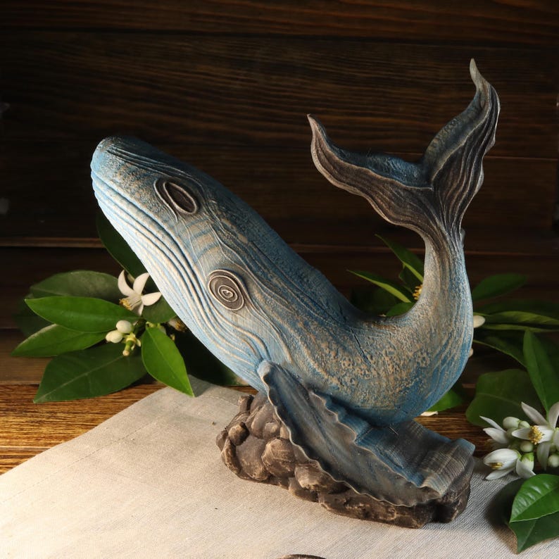 An opposite side view of the hand-carved wood blue whale figurine, showing the textured sea animal sculpture facing left with its tail arched upward. The ocean decor sculpture features detailed blue textures on a dark rock-like base and is available with a personalization option.